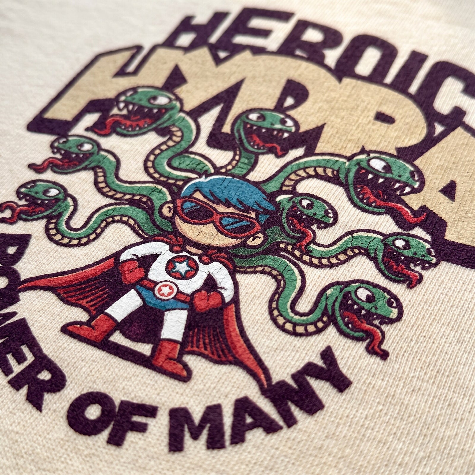 Macro de l'imprime Heroic Hydra, petit super-heros a cape rouge et serpents verts, sur du coton beige