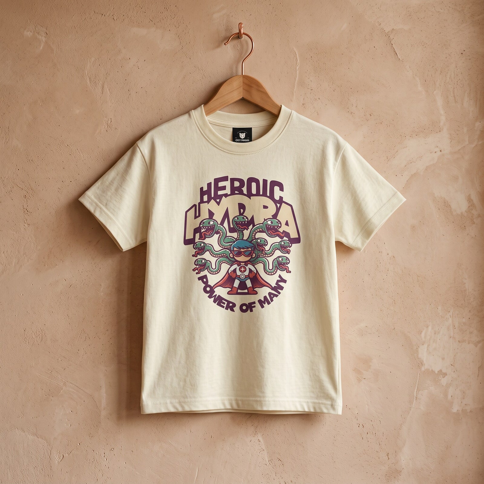 T-shirt enfant beige imprime super-heros et hydre aux tetes de serpents, sur cintre devant un mur terracotta