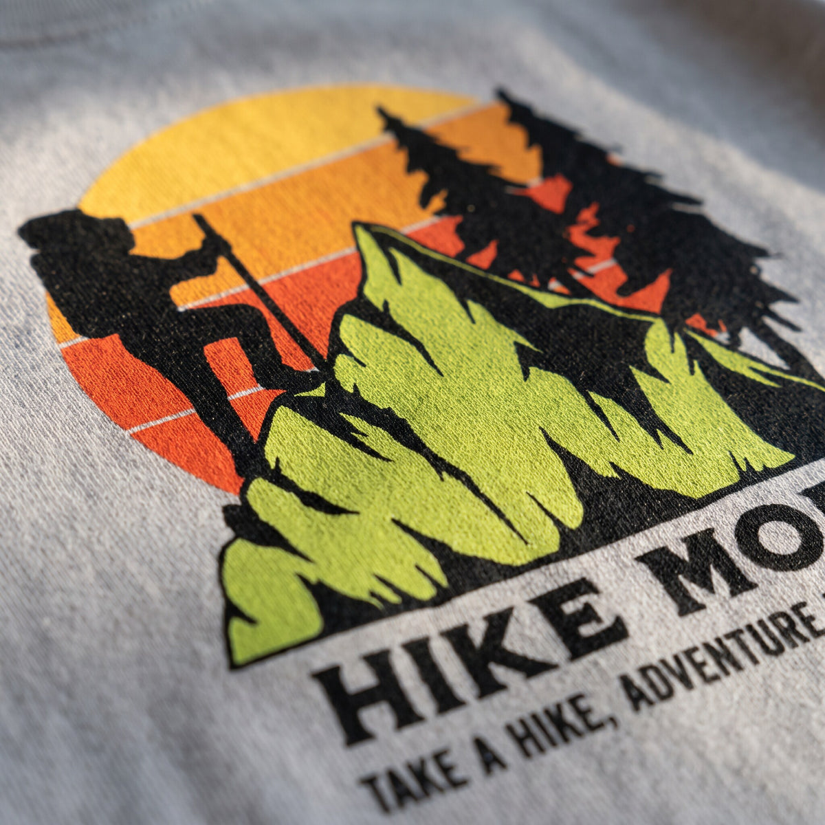 Gros plan sur l'imprime Hike More, randonneur en ombre chinoise devant un soleil degrade orange et jaune