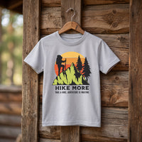 T-shirt enfant gris clair Hike More, randonneur et soleil couchant retro, sur cintre devant une cabane en bois
