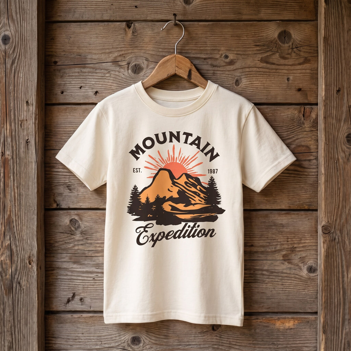 T-shirt enfant ecru sommet et sapins Est 1987, suspendu a un cintre en bois sur un mur de planches brutes