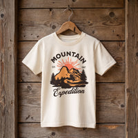 T-shirt enfant ecru sommet et sapins Est 1987, suspendu a un cintre en bois sur un mur de planches brutes
