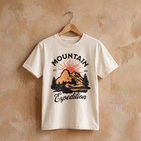 T-shirt enfant ecru Mountain Expedition, montagne et soleil levant, sur cintre devant un mur de platre beige