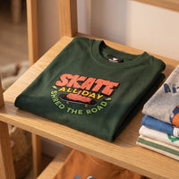 T-shirt enfant vert fonce Skate All Day plie sur une etagere en bois clair a cote d'une pile de t-shirts