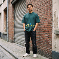 T-shirt oversized vert sapin imprimé fleurs cartoon, porté dans une ruelle pavée devant un mur de briques rouges