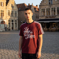 Homme en t-shirt bordeaux rétro Le pays de la Bière sur une place pavée aux façades flamandes, lumière de fin de journée