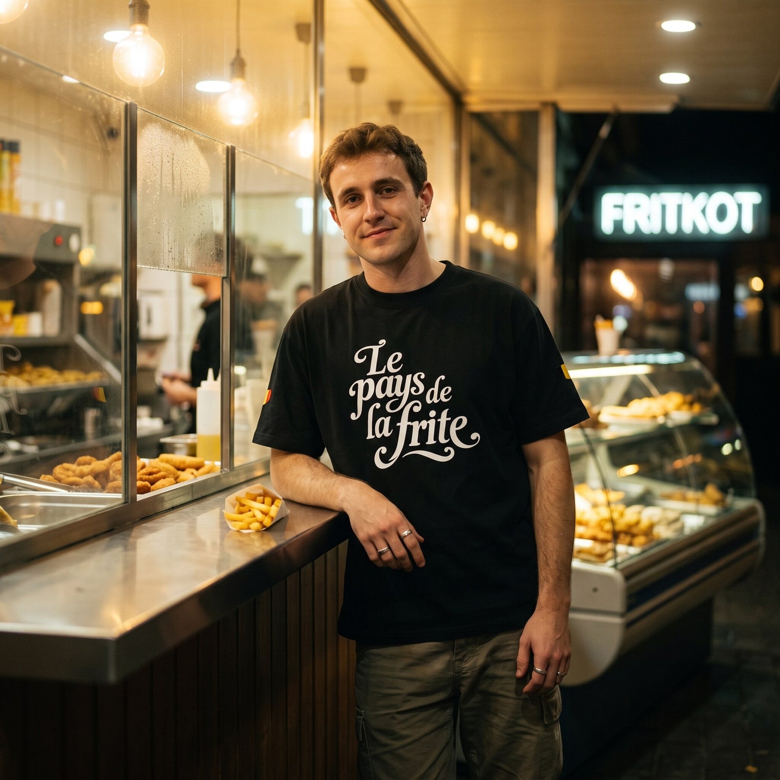 T-shirt oversized noir Le pays de la frite, porté accoudé au comptoir d'une friterie belge sous l'enseigne Fritkot
