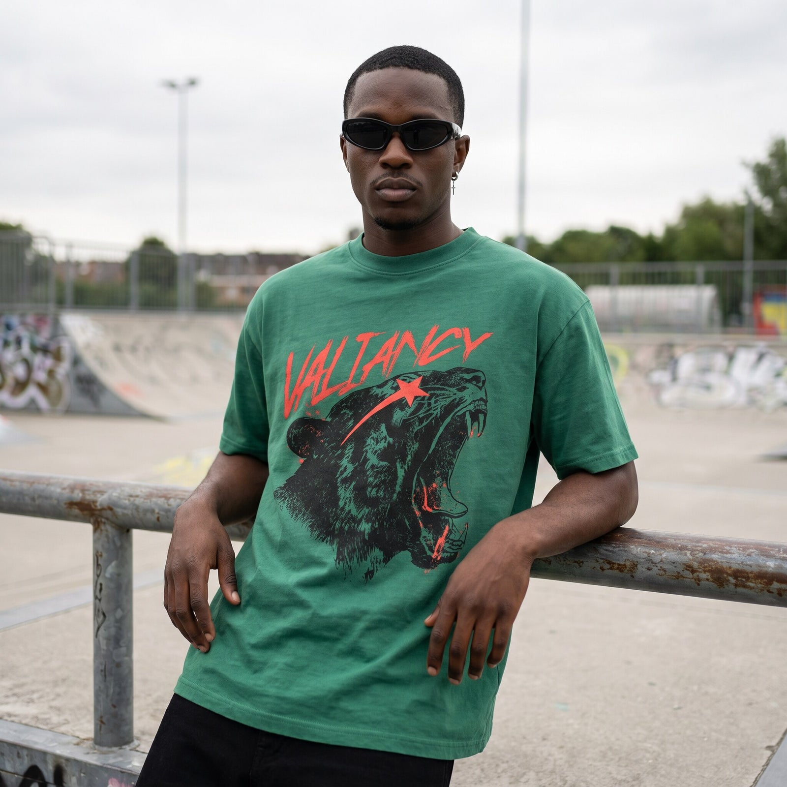 Homme en t-shirt vert Valiancy adosse a une rambarde rouillee devant les rampes graffitees d'un skatepark