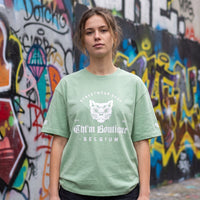 Femme en t-shirt vert amande imprimé tête de chat gothique et lettrage blanc, devant un mur de graffitis colorés