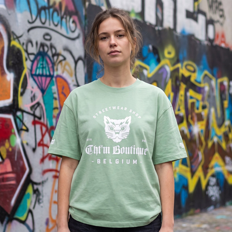 Femme en t-shirt vert amande imprimé tête de chat gothique et lettrage blanc, devant un mur de graffitis colorés