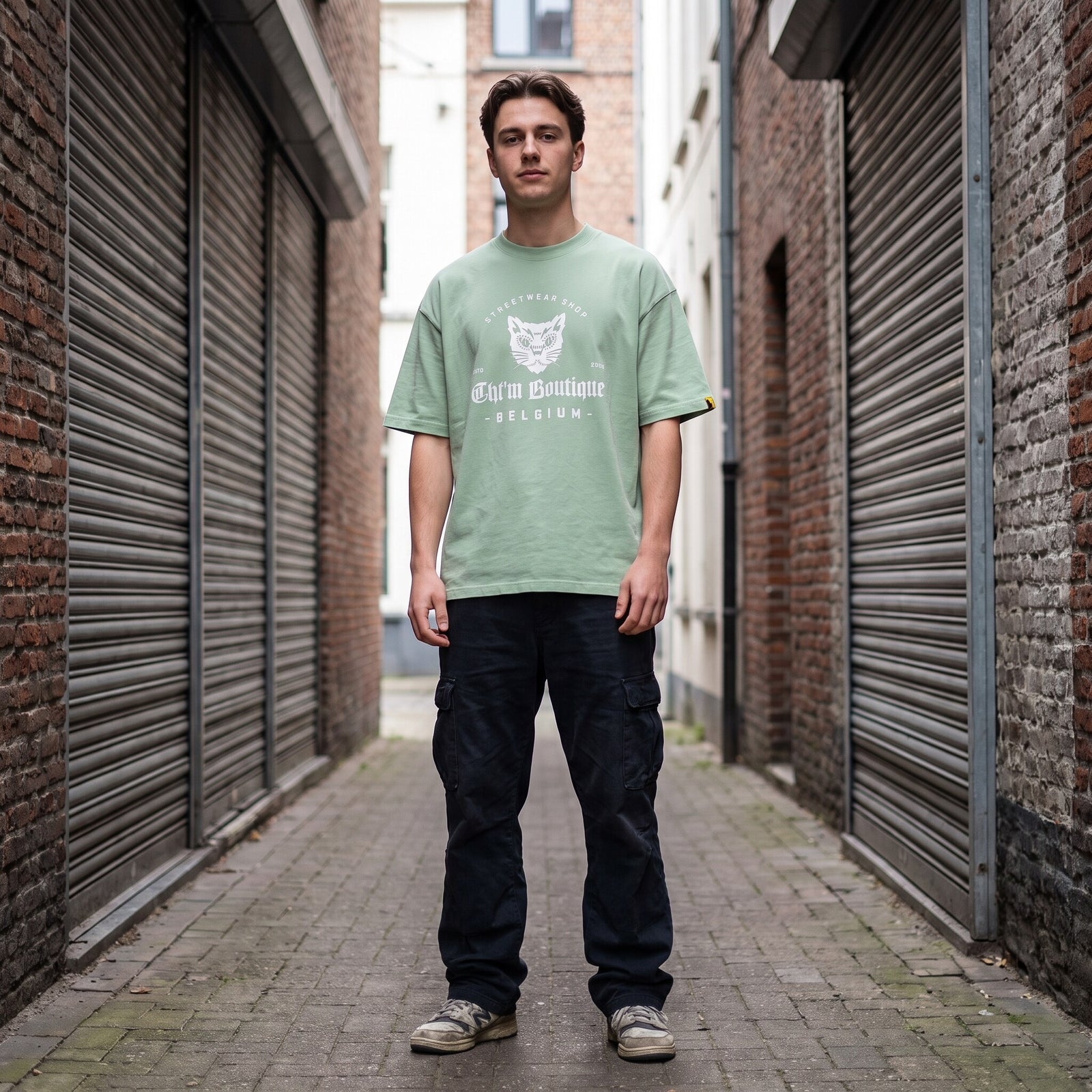 T-shirt oversized vert amande au logo chat gothique CHT'M Boutique Belgium, porté dans une ruelle pavée aux rideaux métalliques