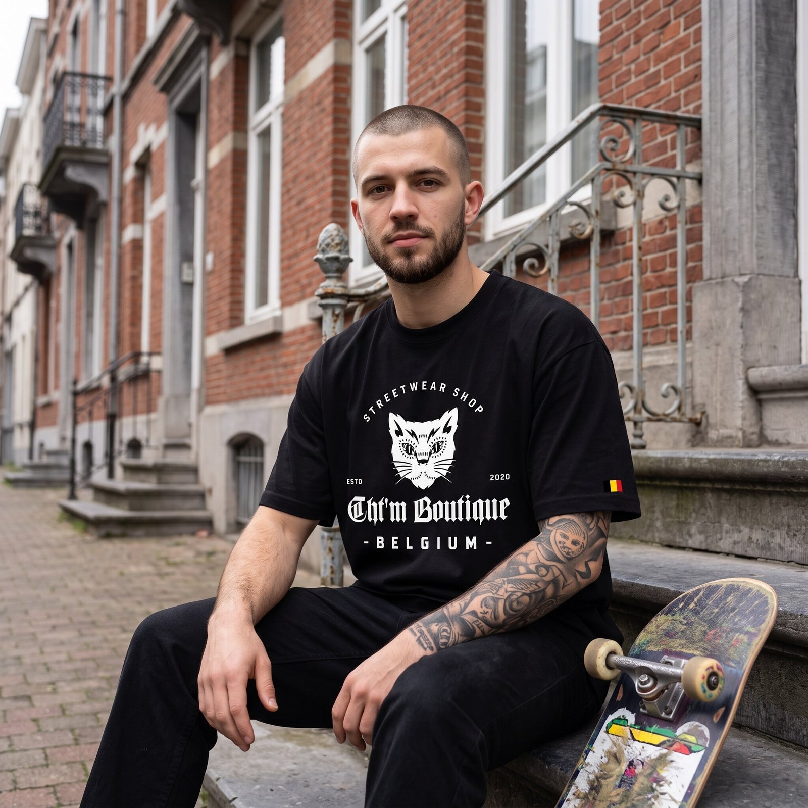 T-shirt oversized noir au logo chat blanc, porté assis sur les marches d'une maison en briques à Bruxelles