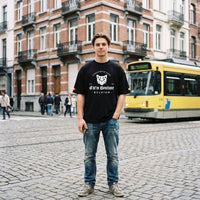 T-shirt oversized noir logo chat streetwear, porté debout sur des pavés devant un tram jaune bruxellois