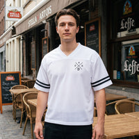 Maillot de sport blanc en mesh, bandes noires aux manches, porte devant une terrasse en rotin d'un cafe belge