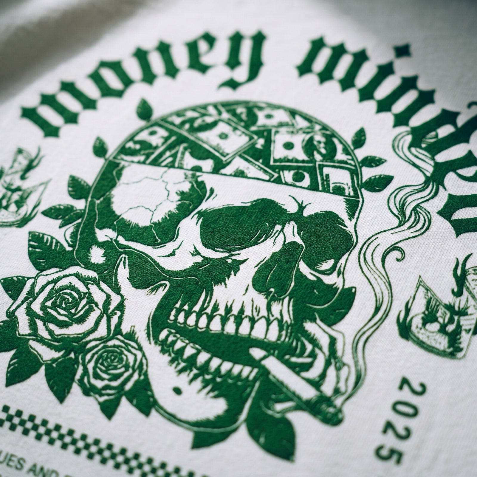 Gros plan sur l'impression verte du crane aux billets et aux roses Money Minded sur le coton blanc du t-shirt