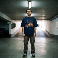 T-shirt oversized bleu marine au badge montagne orange Adventure, porté sur la rampe d'un parking souterrain