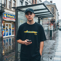 T-shirt noir a l'imprime cartoon jaune, porte devant l'enseigne d'une friterie et un abribus sous la pluie