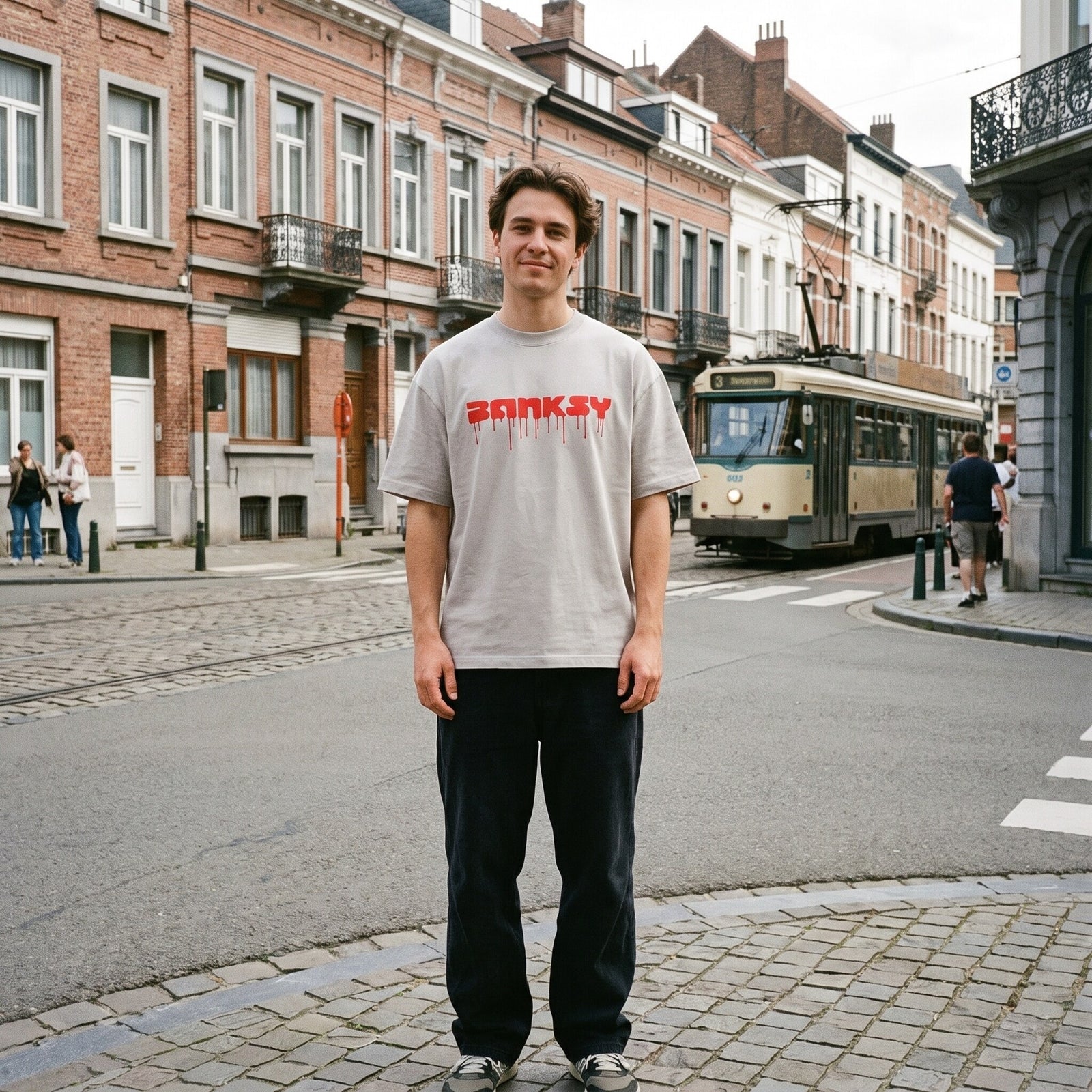 T-shirt oversized blanc cassé imprimé Banksy rouge, porté en plein pied à un carrefour pavé, tram bruxellois en fond