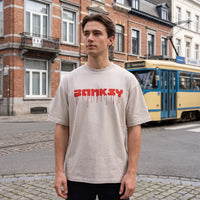 T-shirt oversized Banksy blanc cassé, lettrage rouge dégoulinant, porté devant un tram jaune et bleu à Bruxelles
