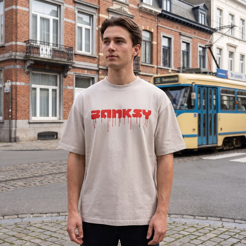 T-shirt oversized Banksy blanc cassé, lettrage rouge dégoulinant, porté devant un tram jaune et bleu à Bruxelles