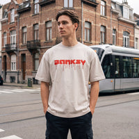 T-shirt oversized Banksy beige, mains dans les poches devant une façade en brique et un tram moderne blanc