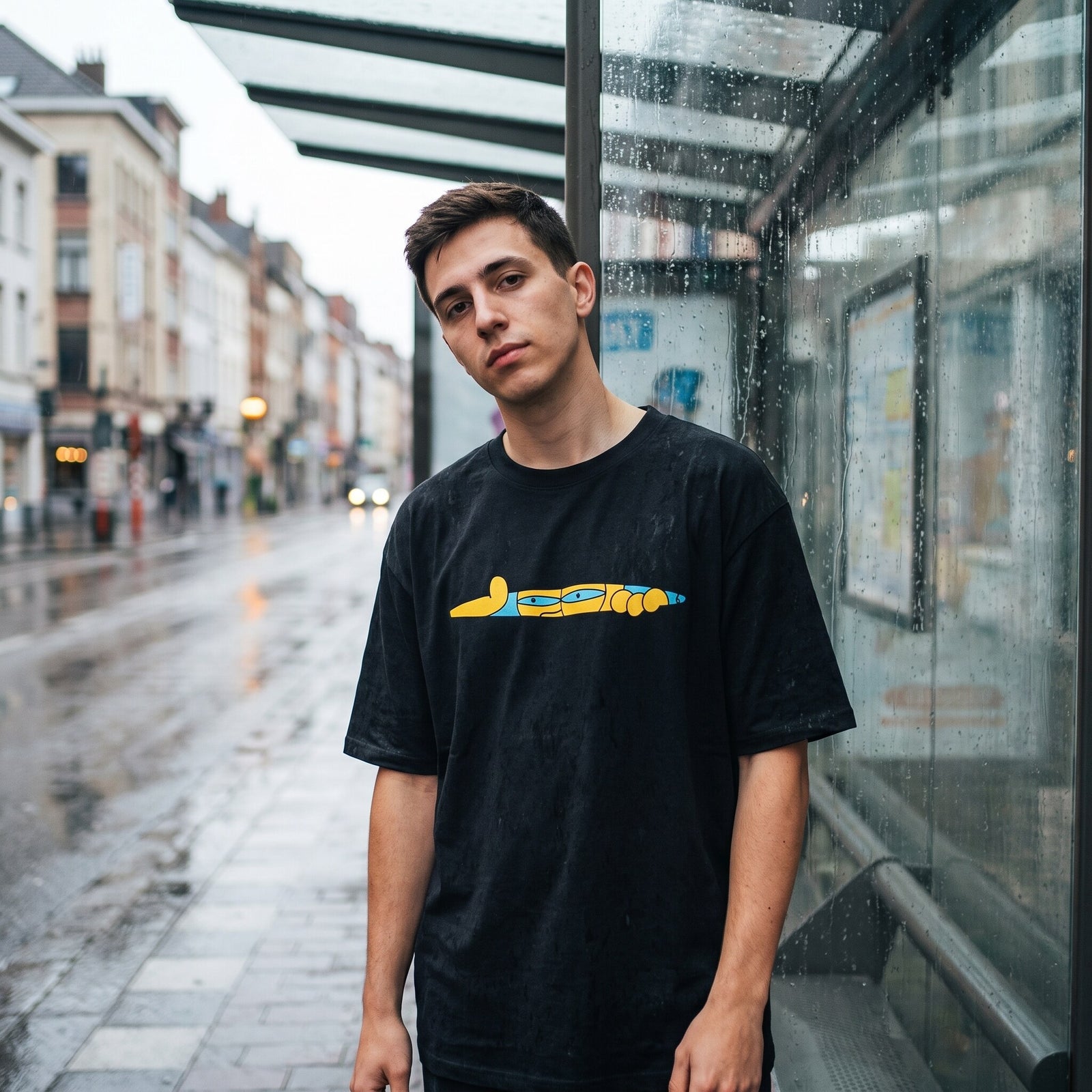 T-shirt oversized noir a bande cartoon jaune et bleue, porte sous un abribus vitre par temps de pluie