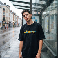 T-shirt oversized noir a bande cartoon jaune et bleue, porte sous un abribus vitre par temps de pluie