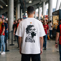 T-shirt oversized blanc, portrait de Doku imprime au dos, porte dans une coursive de stade parmi les supporters