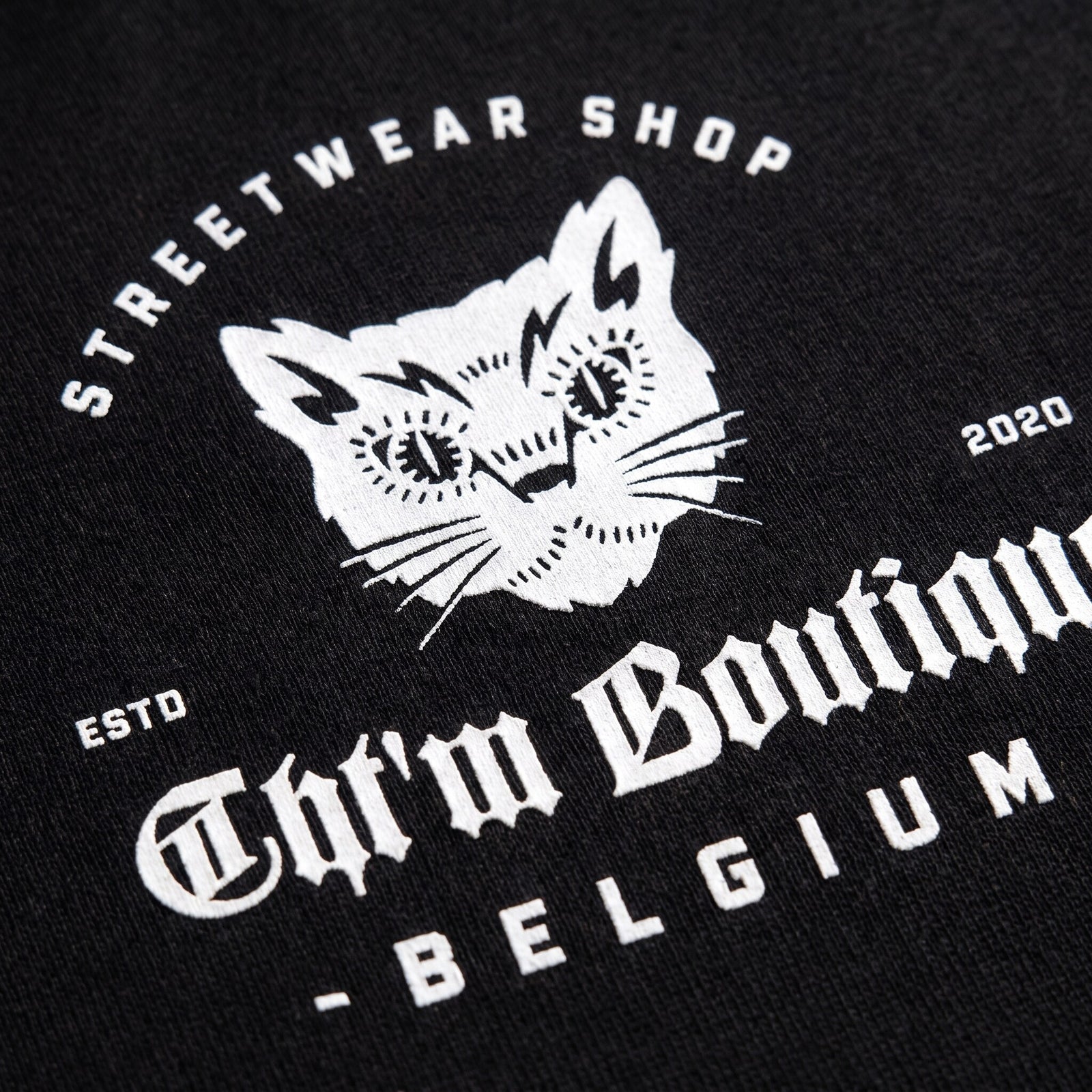 Gros plan sur la tete de chat blanche et le lettrage Streetwear Shop Belgium imprimes sur du coton noir