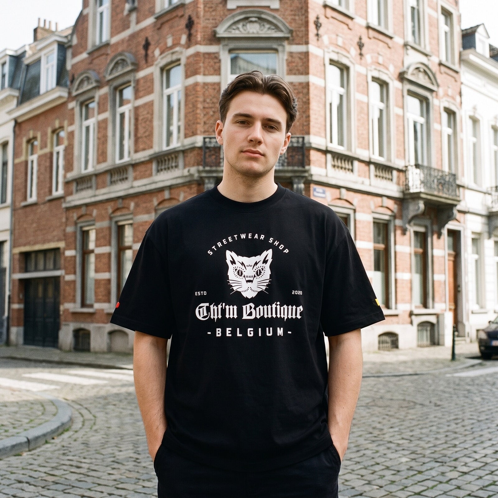 T-shirt oversized noir au logo chat blanc CHT'M Boutique Belgium, porte a un coin de rue pavee bruxelloise