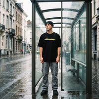 Silhouette entiere en t-shirt noir cartoon et pantalon cargo gris, debout dans un abribus sur des paves mouilles