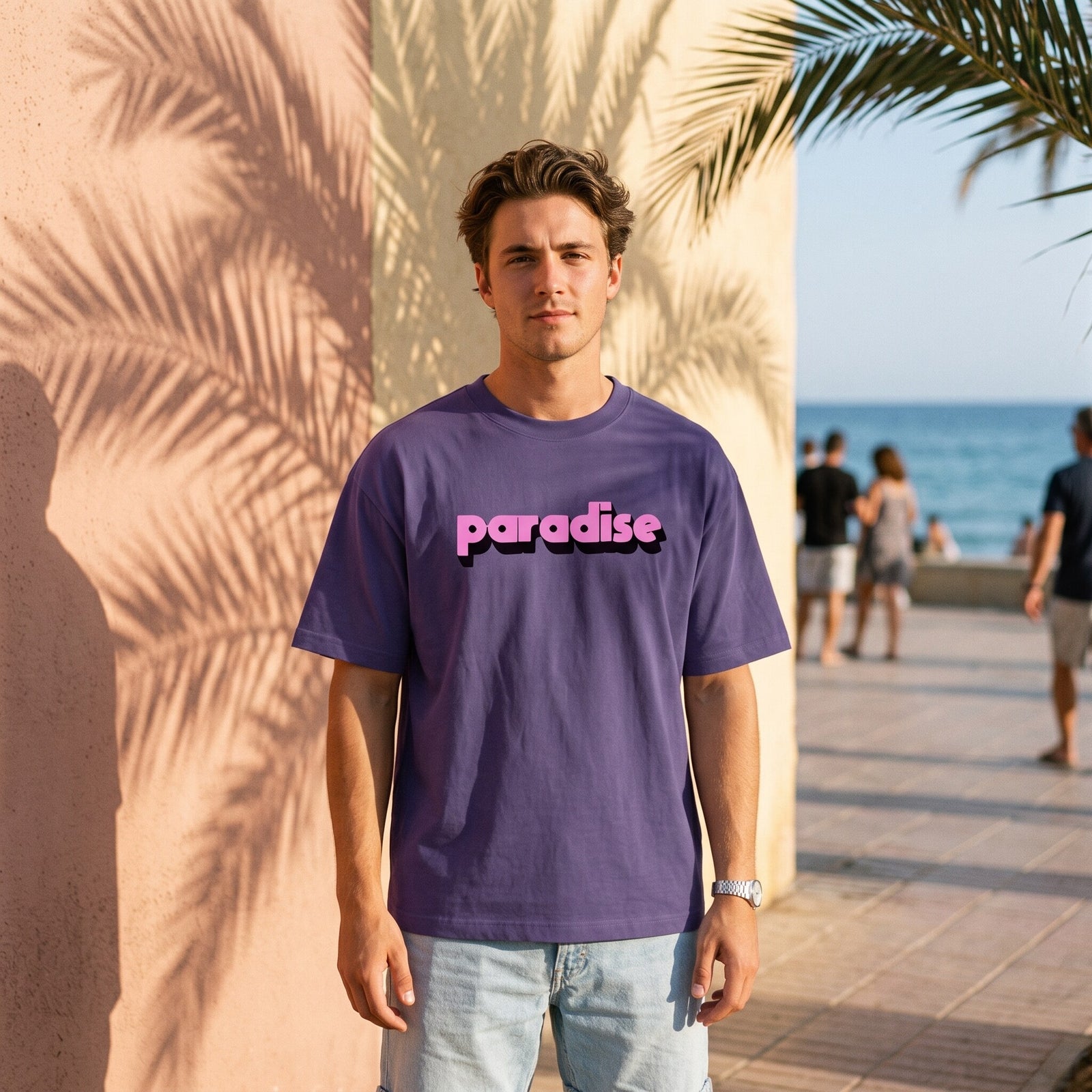 T-shirt oversized violet brume paradise, porte sur une promenade en bord de mer sous l'ombre des palmiers