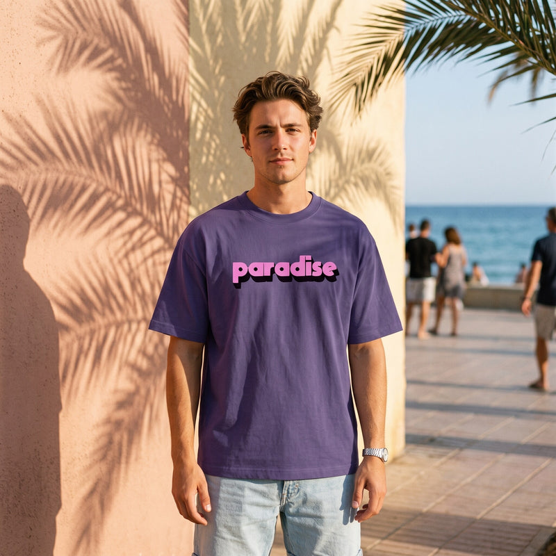 T-shirt oversized violet brume paradise, porte sur une promenade en bord de mer sous l'ombre des palmiers