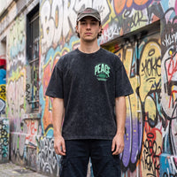 Homme en casquette et t-shirt oversized snow wash noir CHT'M, petit logo PEACE vert, mur de graffitis