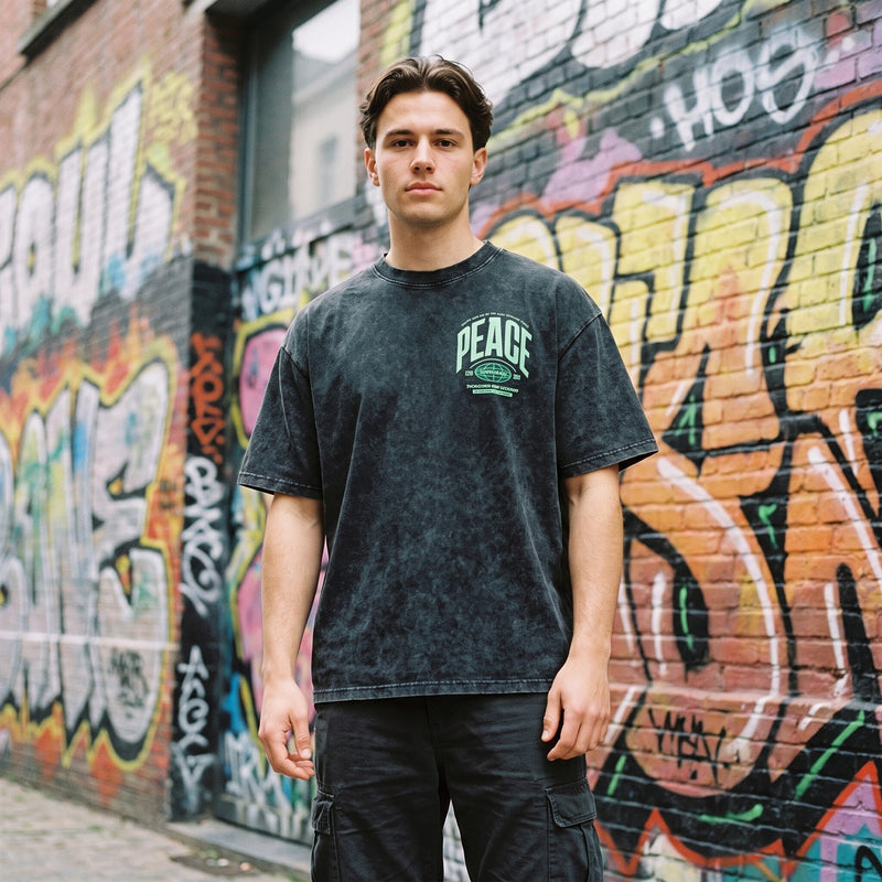 T-shirt oversized noir délavé snow wash, logo PEACE vert menthe, porté dans une ruelle couverte de graffitis