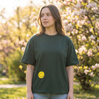 Femme en t-shirt oversized vert sapin à smiley jaune, dans un parc au printemps devant un magnolia en fleurs