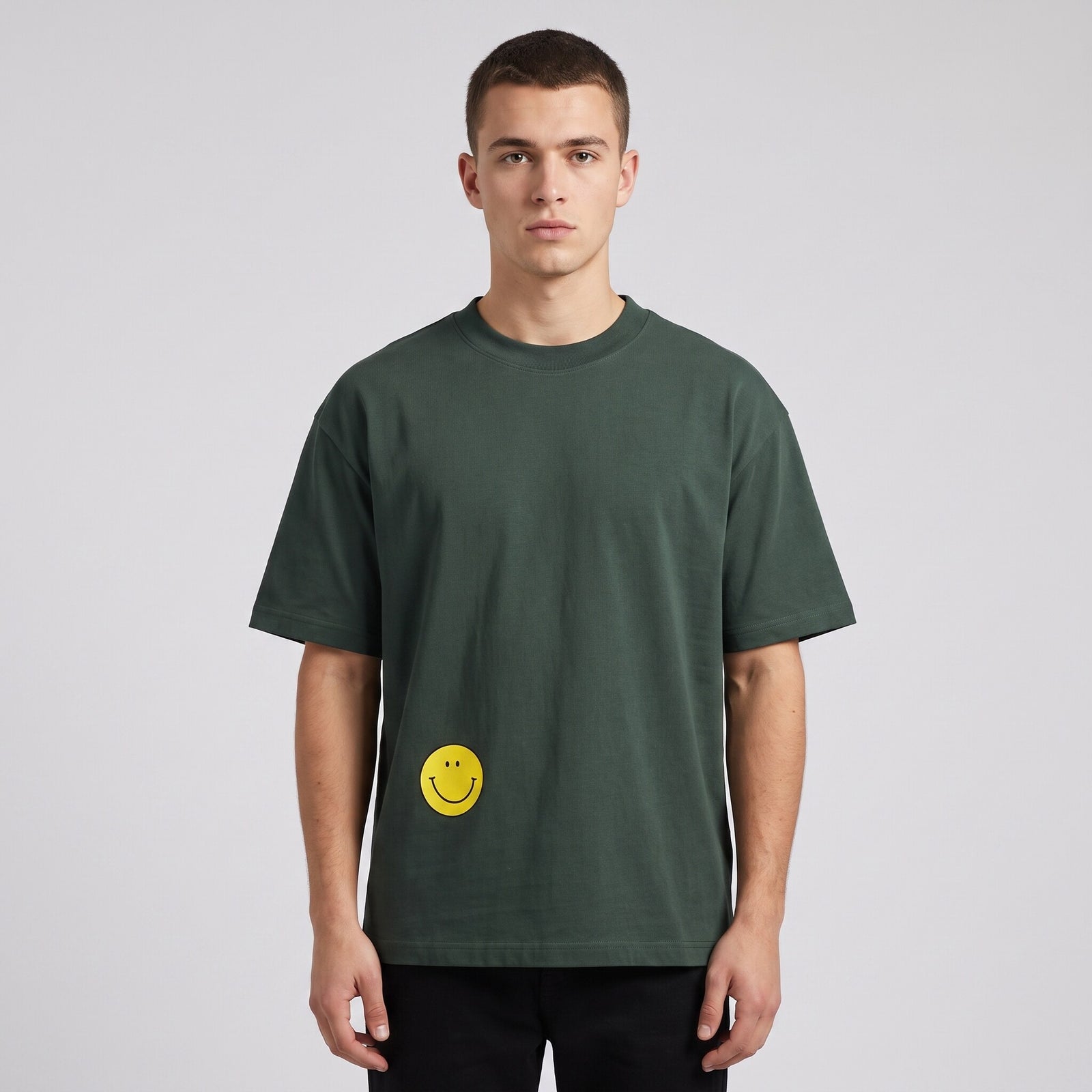T-shirt oversized vert sapin avec smiley jaune imprimé en bas à gauche, porté en studio sur fond gris clair
