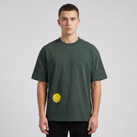 T-shirt oversized vert sapin avec smiley jaune imprimé en bas à gauche, porté en studio sur fond gris clair