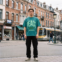 T-shirt vert delave PEACE porte en pied sur une place commercante de Bruxelles, tram de la STIB en arriere-plan
