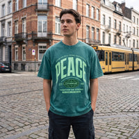 T-shirt oversized PEACE vert sapin, mains dans les poches devant un tram jaune sur les paves bruxellois