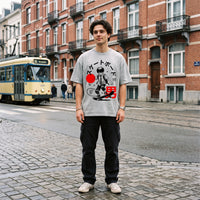 T-shirt gris clair oversized au soleil rouge et skateur manga, porté au coin d'une rue pavée où passe un vieux tram