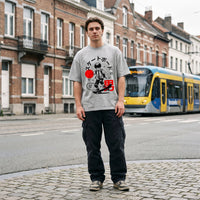 Coupe oversized grise et imprimé skateur manga noir et rouge, portée sur les pavés devant un tram moderne de la STIB