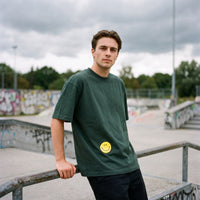 Homme appuyé sur une rambarde de skatepark, t-shirt vert sapin au smiley jaune et rampes de béton taguées