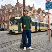 T-shirt oversized vert sapin au petit smiley jaune sur le bas, porté devant un tram bruxellois sur une rue pavée