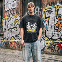 T-shirt noir délavé Stop Wars porté avec un bonnet beige et un jean clair déchiré devant une fresque de street art