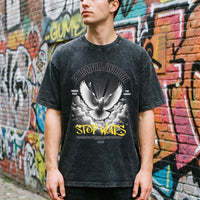 T-shirt oversized noir délavé à colombe de la paix, porté dans une ruelle bordée de briques et de tags colorés
