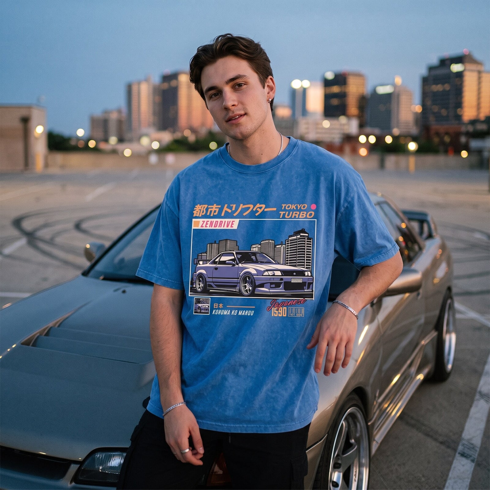 T-shirt oversized bleu Tokyo Turbo, porté adossé à une voiture de sport sur un parking en toit-terrasse au crépuscule
